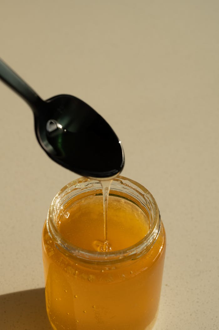 A close-up of golden honey dripping from a black spoon into a jar, captured in bright lighting.
